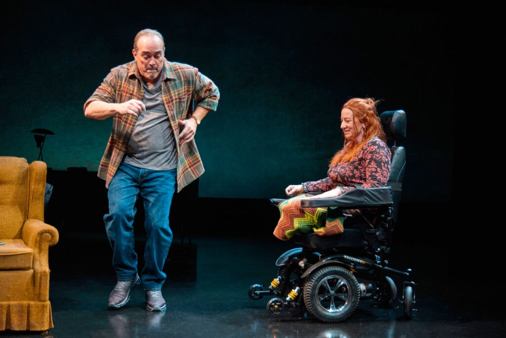 scene from a play stage showing a woman in a power chair watching a man in flannel shirt dancing