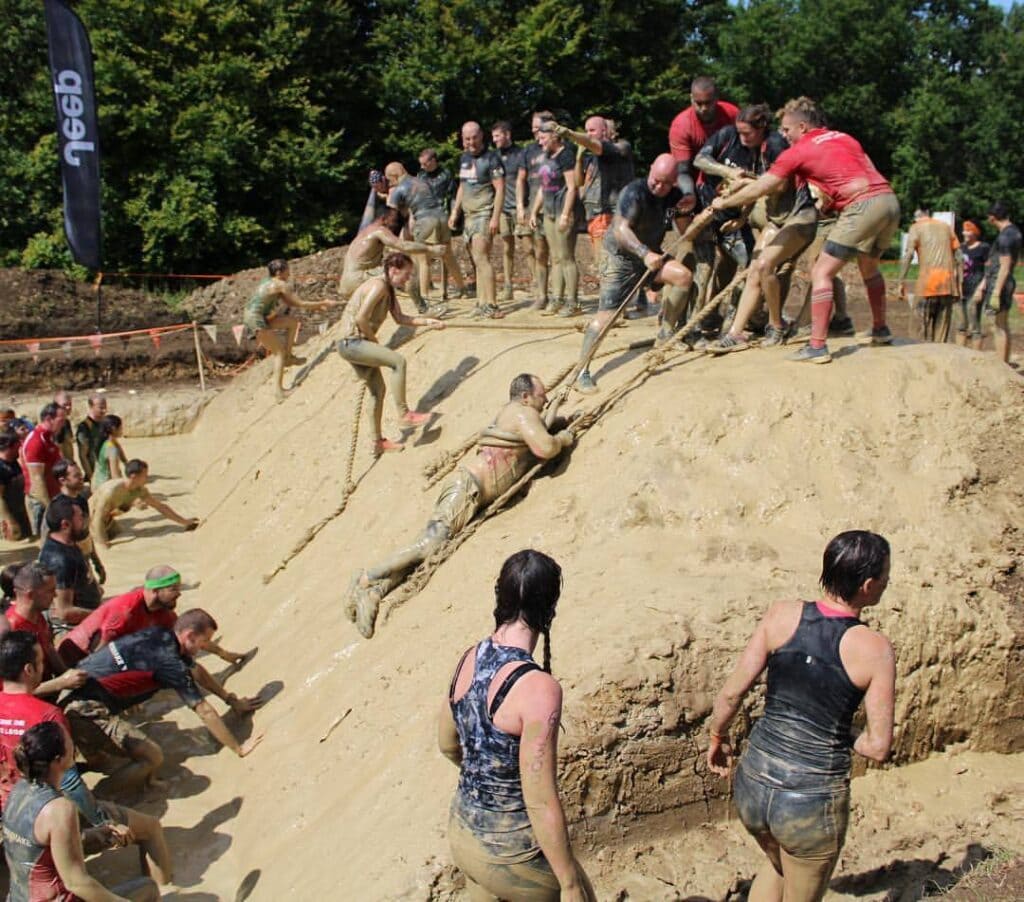 Muddy bank with dozens of athletes at top and bottom. In foreground a white man, Andrew Holley, is holding onto a rope as teammates pull him up the bank. 