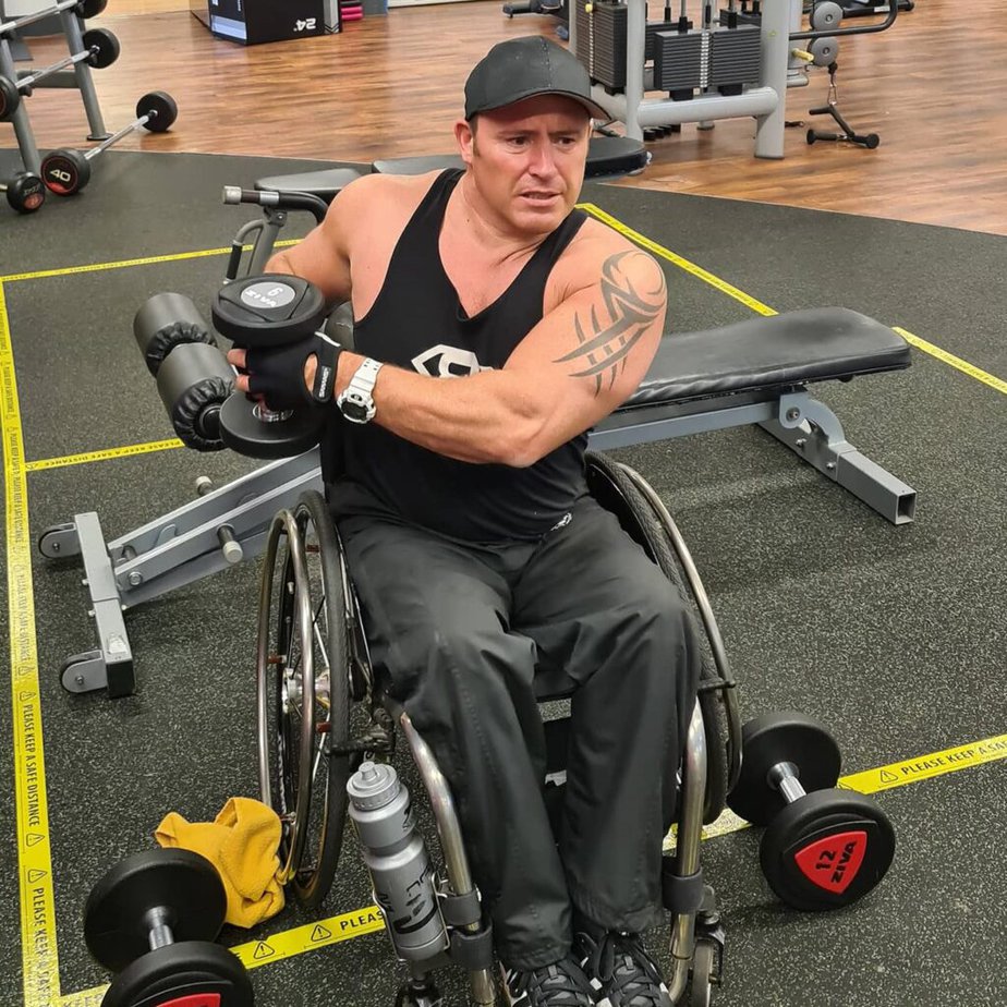White male manual wheelchair user working out with a dumbbell at a fitness center. 