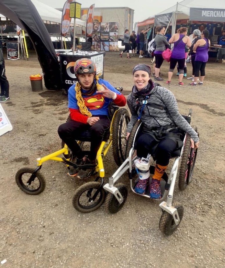 David Tyson Perry and Jesi Stracham, both using off-road wheelchairs sit by by side smiling at camera. Event tents are in background.