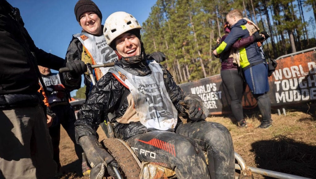 White female wheelchair user smiling in muddy field. Her wheels are covered in mud. She is smiling, wearing a race bib and helmet, and being pushed by a teammate. 