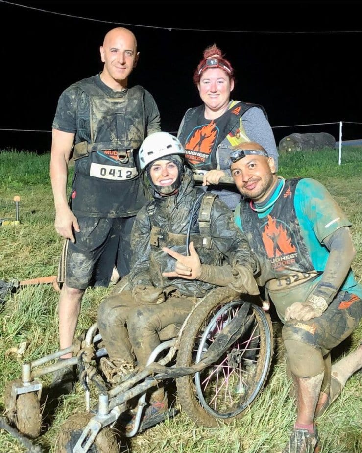 White female wheelchair user, covered in mud, using off-road wheelchair, surrounded by smiling teammates. 