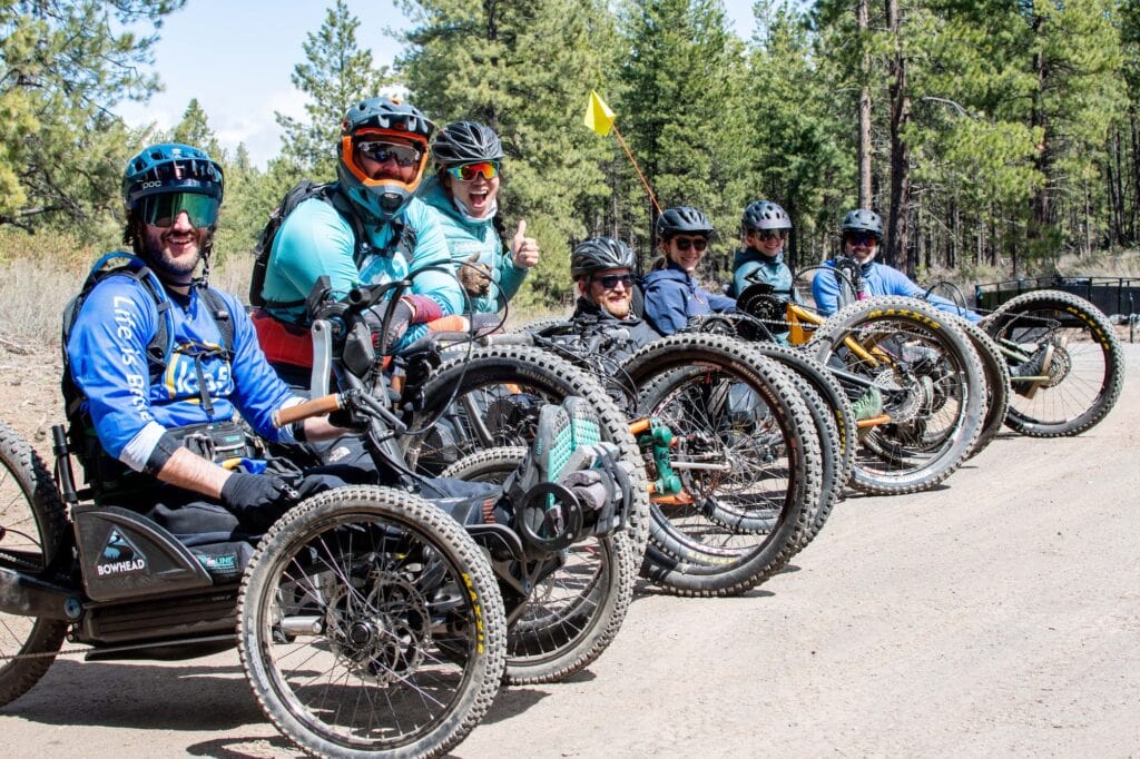 Line of people sitting on adaptive mountain bikes of various styles. Everyone is wearing helmets and smiling at the camera. 