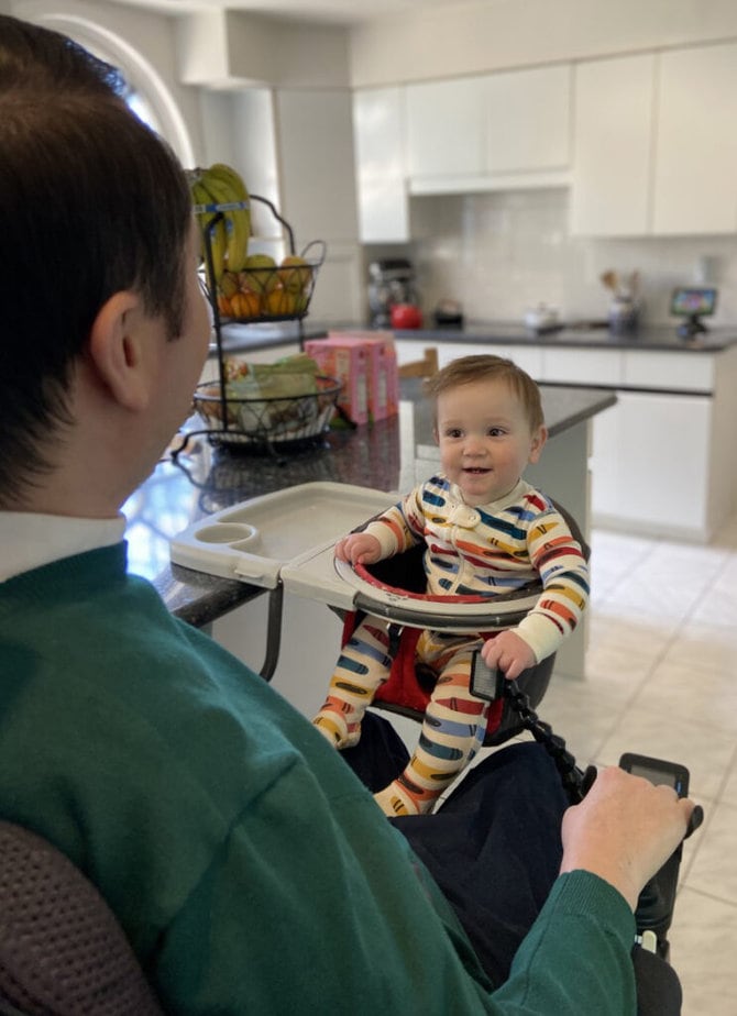 Rear view of man in powerchair looking at baby in a high chair smiling back at him