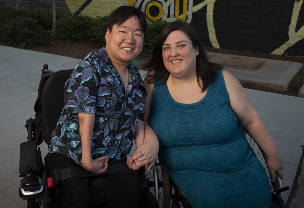man and woman wheelchairs users closely seated next to each other holding hands