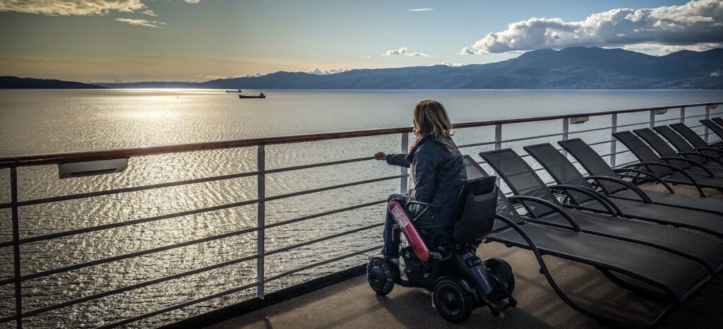 White woman using a power wheelchair, sits on deck of cruise ship looking out at sea with mountains in the background.