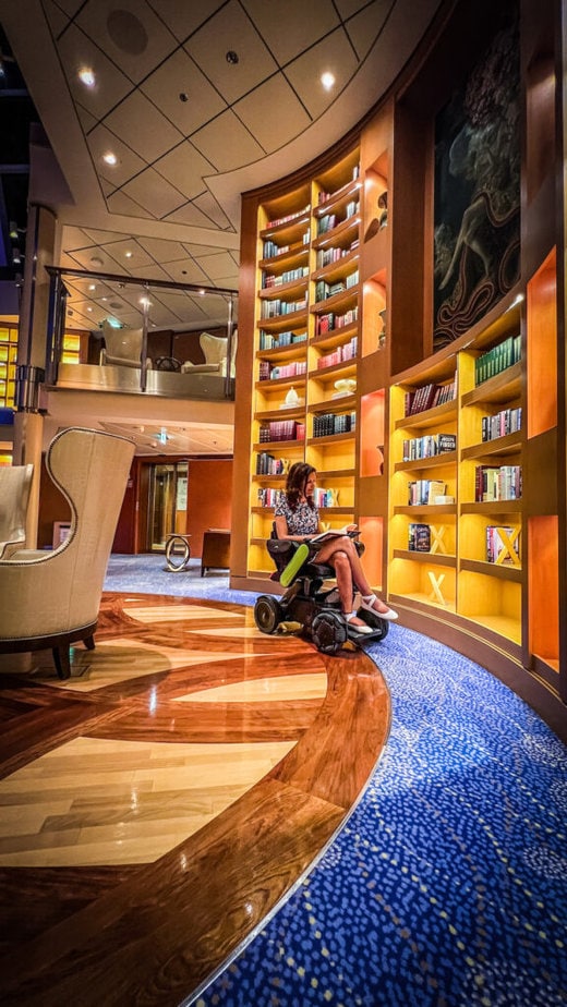 Sylvia Longmire, a white woman using  a power wheelchair, sits reading a book next of a tall bookshelf in a luxurious cruise ship.