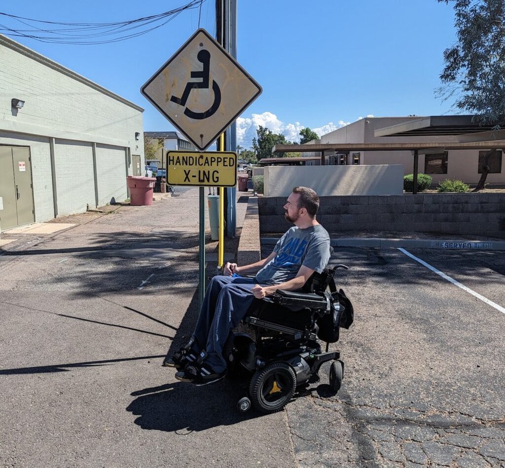 White man using power wheelchair, looking up at street sign with "handicapped crossing" set in parking lot