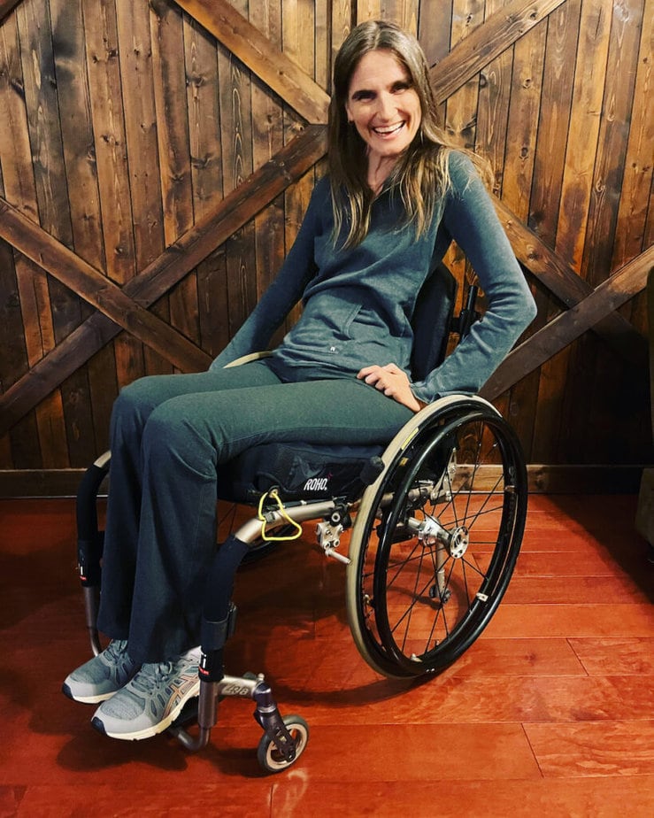 White woman with long, straight brown hair using manual wheelchair and smiling at camera.