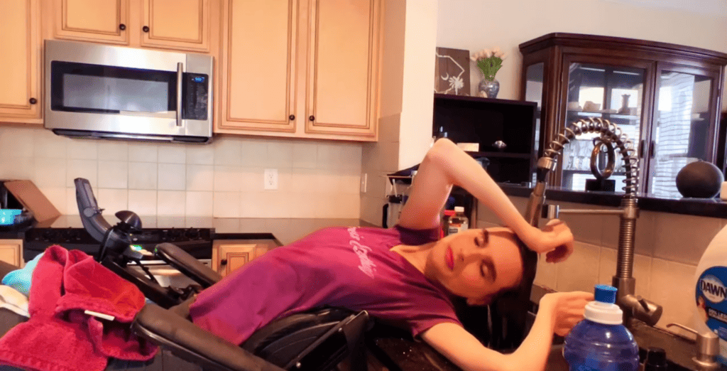 A white woman lays back in her power wheelchair with her hair over a kitchen sink.