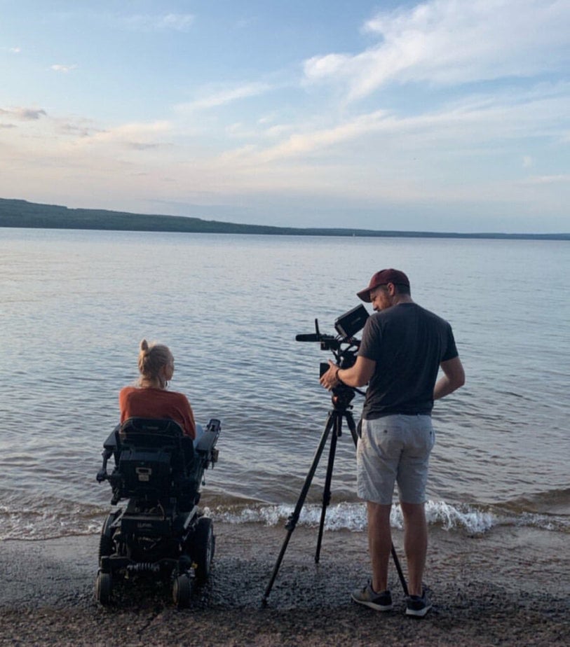 woman in powerchair at waters edge on the beach being filmed by  cameraman