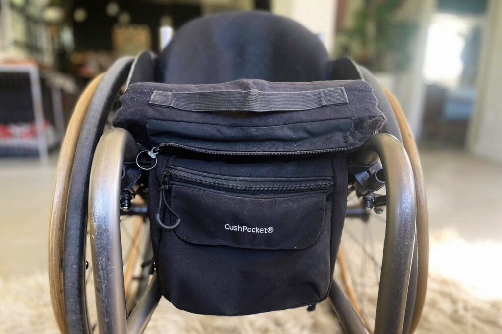 Closeup of front of manual wheelchair with black bag hanging below black cushion. Bag has a zipper and "CushPocket" in white lettering on front flap.
