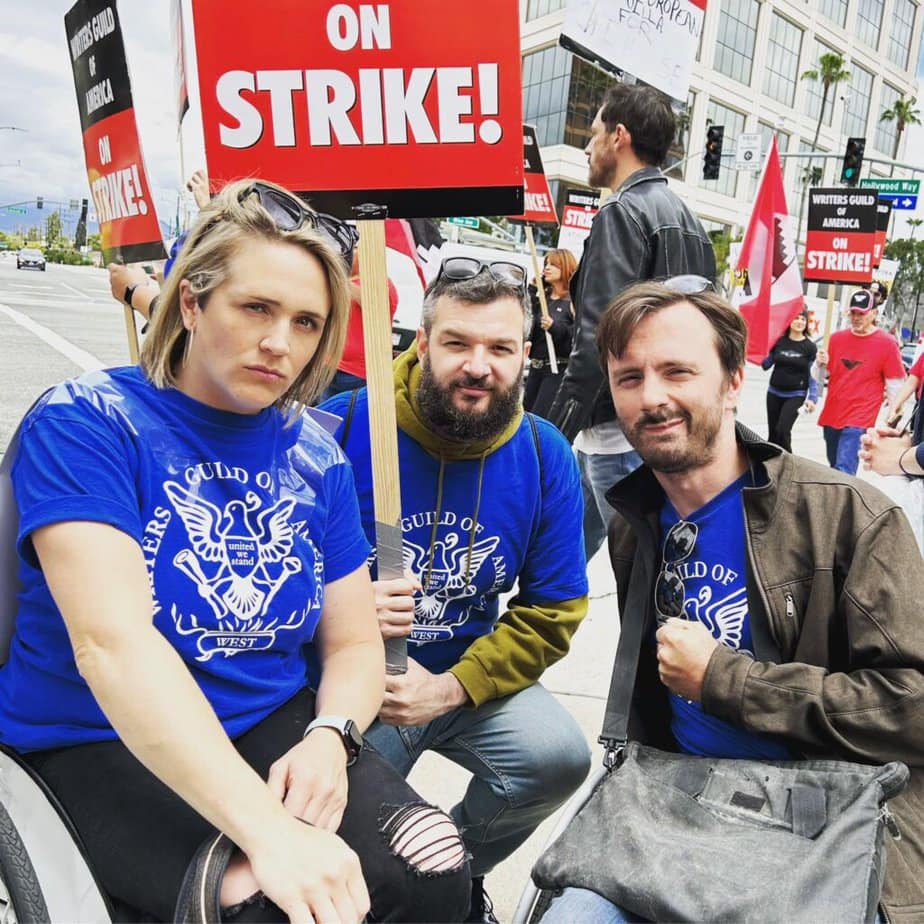 Two wheelchair users, a woman on the left and a man on the right, with a nondisabled man in between look at the camera. The nondisabled man holds a sign that says, "On strike"!