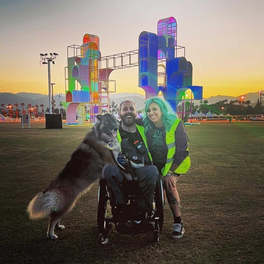 man in wheelchair at an outdoor concert with a dog on his right and woman with green hair standing next to him on his left