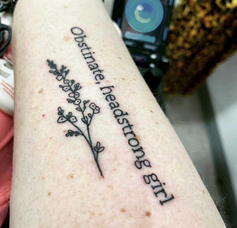 Tattoo on a woman's arm in black ink that reads" Obstinate, headstrong girl" with a branch of flowers below