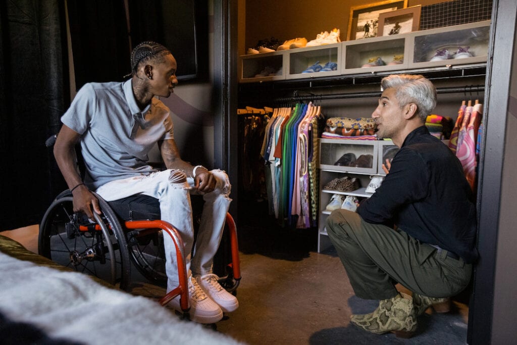 Two men in front a closet full of men's cloths. One left is a young Black man, Ray "Speedy" Walker, using a manual wheelchair, he is wearing a button up shirt and light colored jeans. On right is an older man with gray hair and wearing stylish clothes. He is crouched down and looking at Walker.