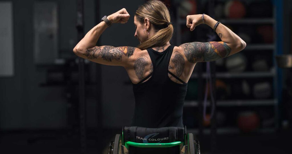 Woman in wheelchair shown from behind with arms raised displaying tattoos on arms, shoulders and back