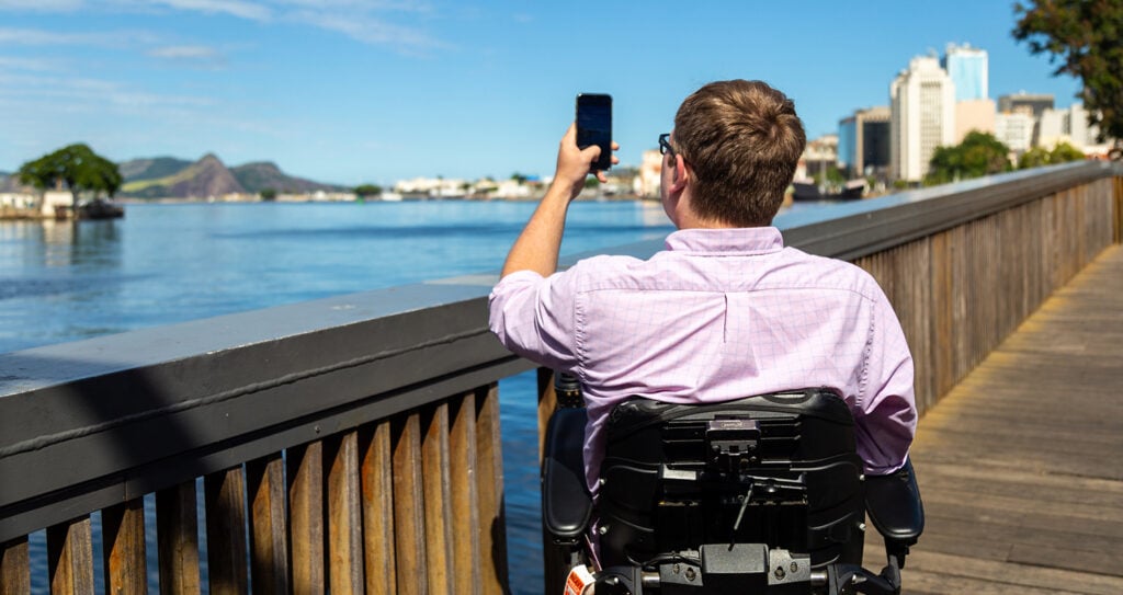 Back view of man in powerchair taking a photo of scenery with water and mountains in background