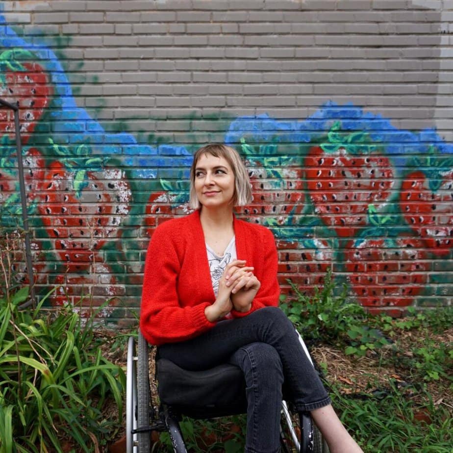 A white woman with blond hair using a manual wheelchair. She is wearing black pants and a red blazer sitting outside in front of a wall with a mural of roses.