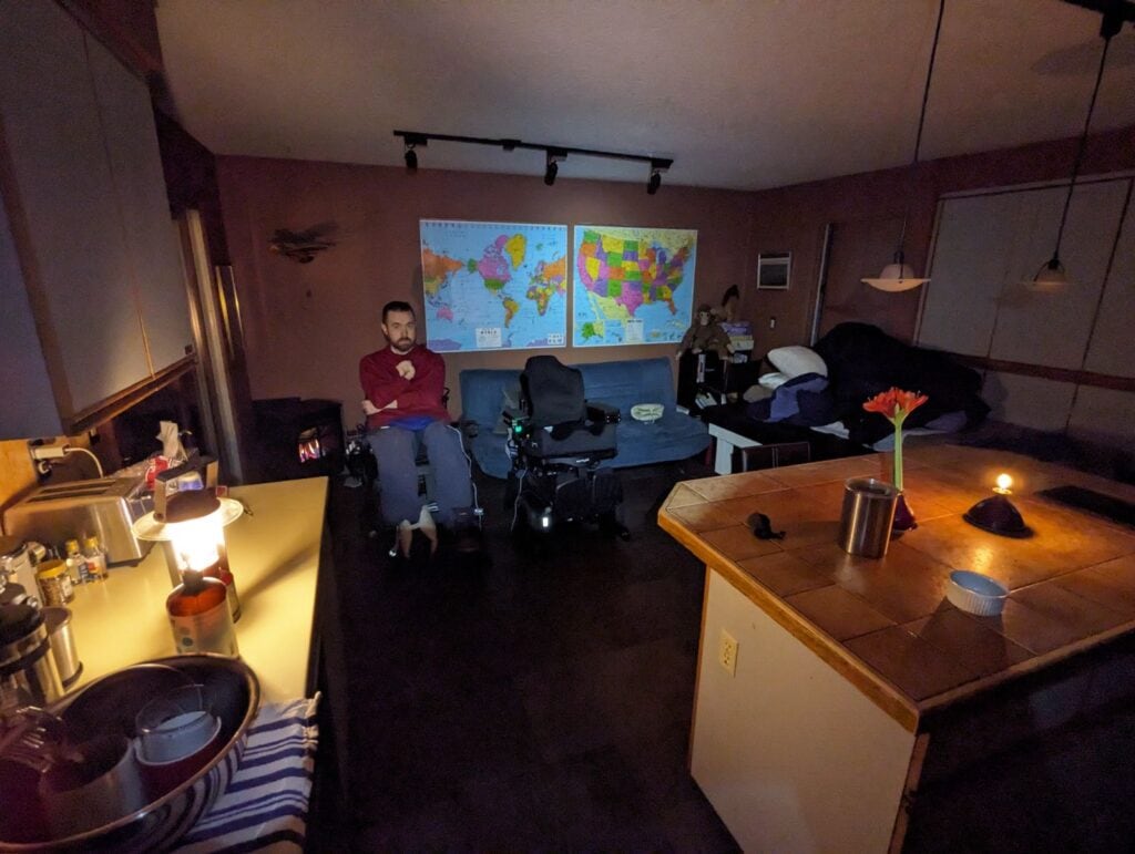 A white in a power wheelchair sits in a dark kitchen, illuminated by laterns on the counter.