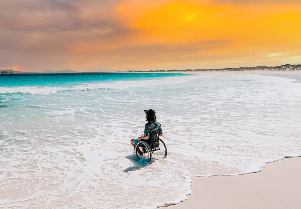 man in wheelchair in surf looking out at ocean with orange sky