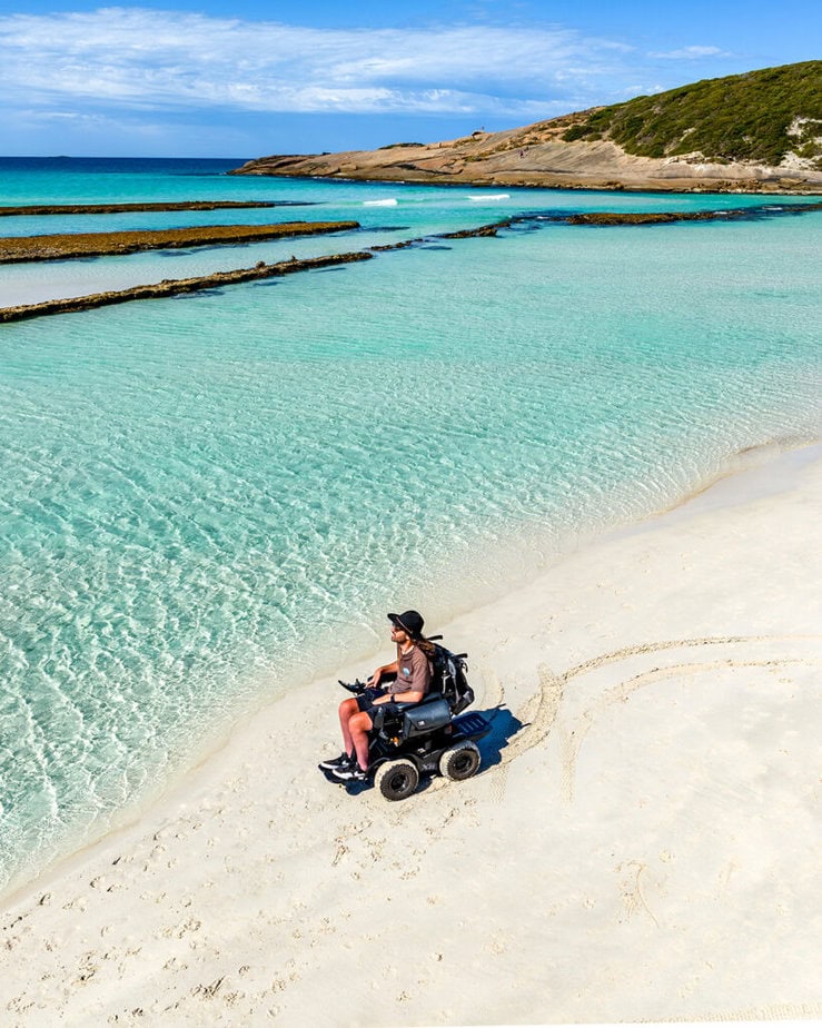 Aerial view of powerchair user riding down white beach with beautiful blue water