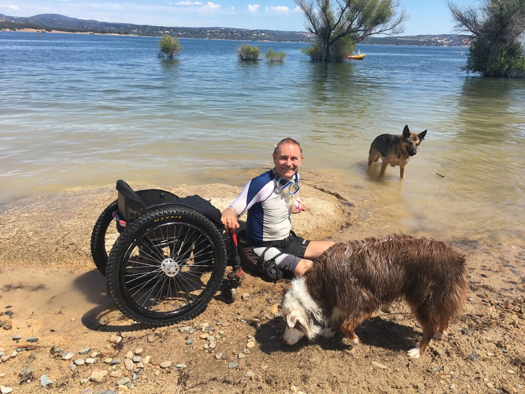 man sitting by waters edge next to wheelchair with two dogs