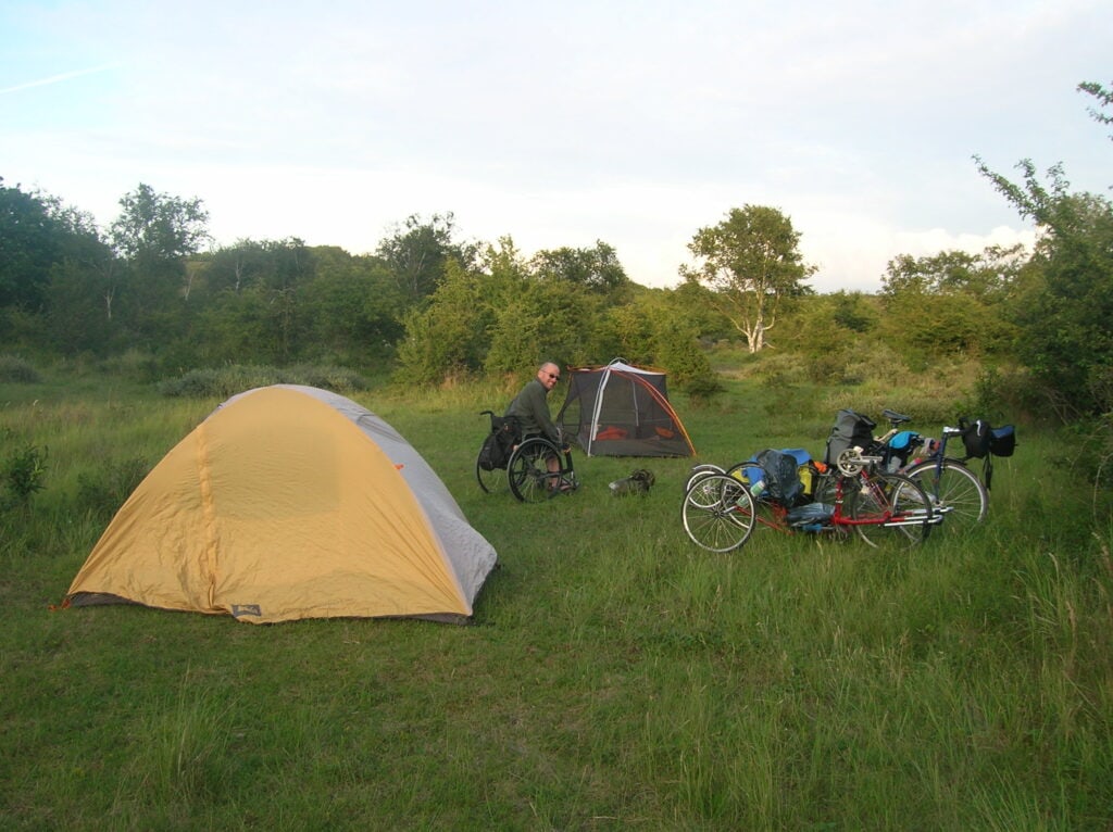 Manual wheelchair user sits between two tents in a grassy field. 