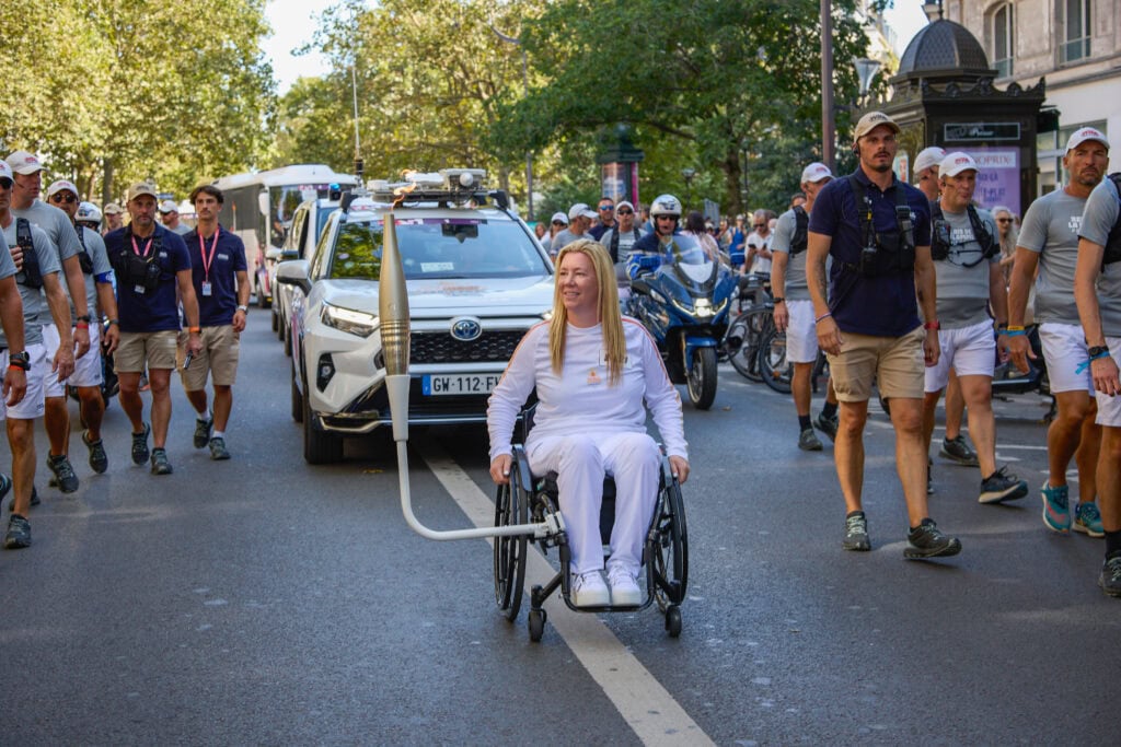 white female manual wheelchair user pushing down a street with crowds around her and the Paralympic torch attached to her chair via a metal arm. 