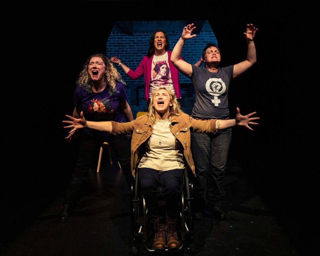 a female manual wheelchair user onstage with arms spread to her sides, singing loudly. three other actors are singing loudly behind her. 