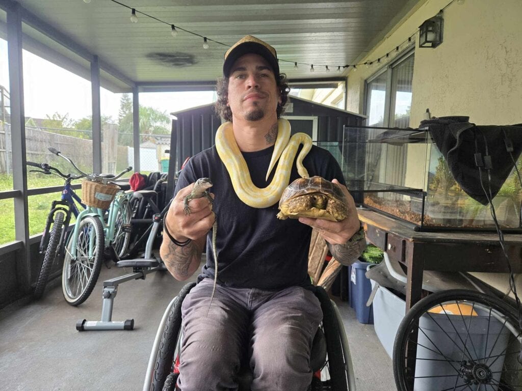 a man using a manual wheelchair sits in a sunroom with a tortoise in one hand, a python around his neck, and a monitor lizard in the other hand.