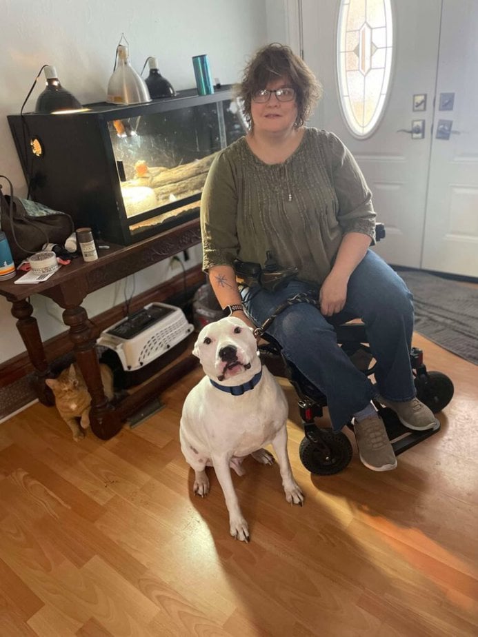 A female power wheelchair user posing inside with a white pit bull-looking dog. 