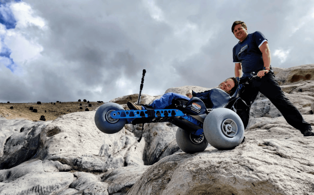 man in an. off-road wheelchair on rocky terrain with another man holding the back handles on the wheelchair to tilt it up in a wheelie. 