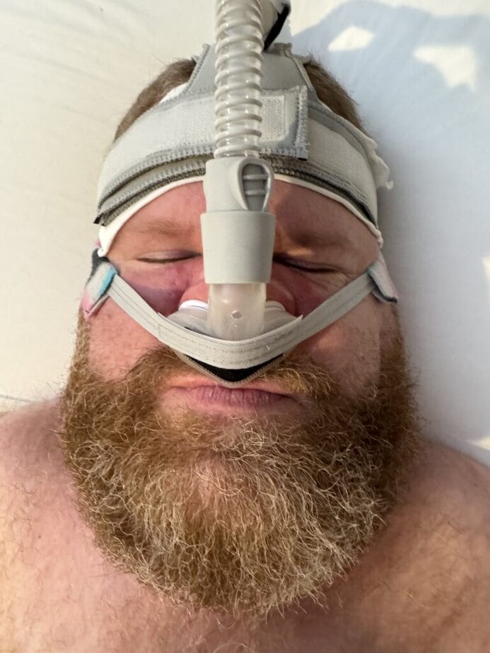 white man with a beard wearing a CPAP mask in bed.