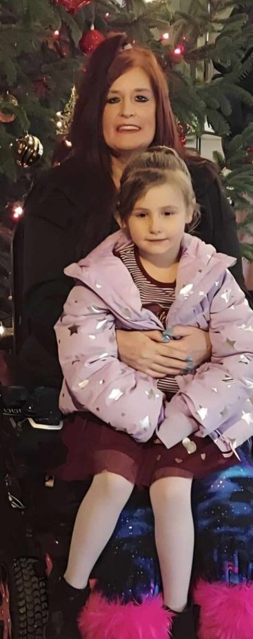 a white woman using a power wheelchair sits with her granddaughter on her lap in front of a Christmas tree.