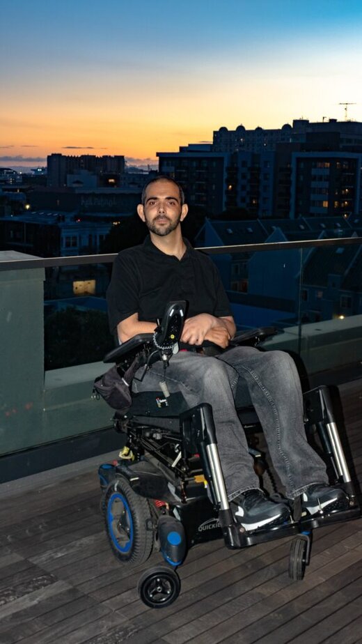 Man using a power wheelchair sitting on a rooftop deck with a city skyline in the background.
