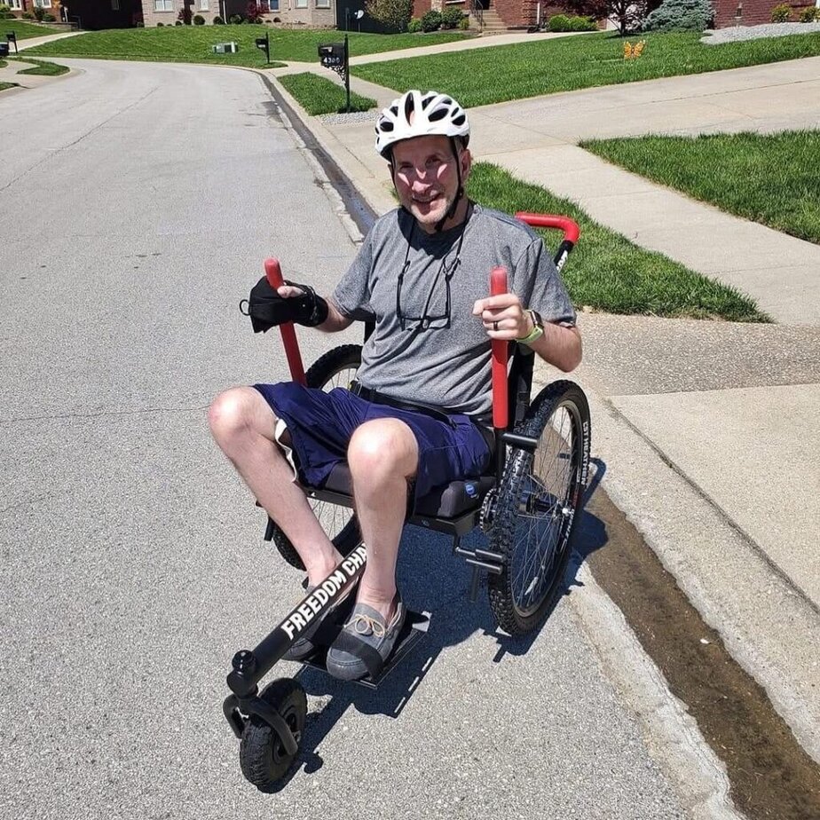 white man sitting in a manual wheelchair with lever handles. He is outside on a neighborhood street and he is wearing a bike helmet. 