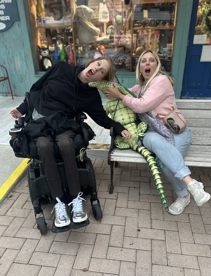 two women, one using a powerwheelchair bending to the side with a silly, surprised look on her face, the oter sitting on a bench holding a toy alligator to the other ones head. 