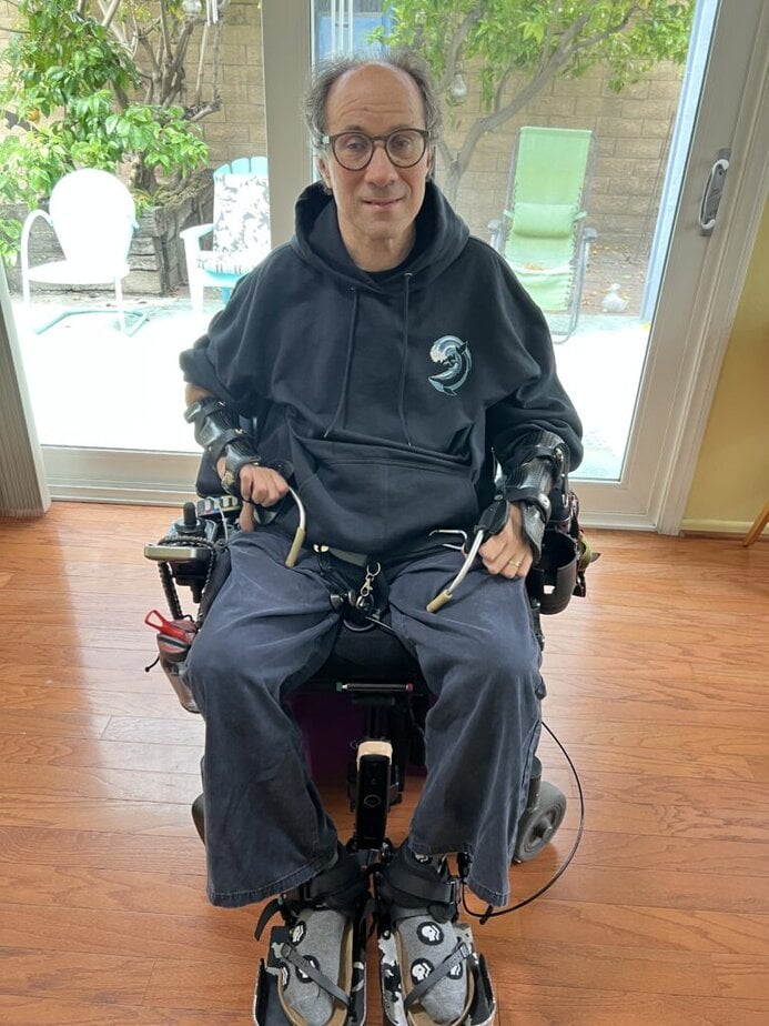 a middle-aged white man using a power wheelchair. He's sitting inside, in front of a french door, wearing a black hoodie and looking at the camera. 