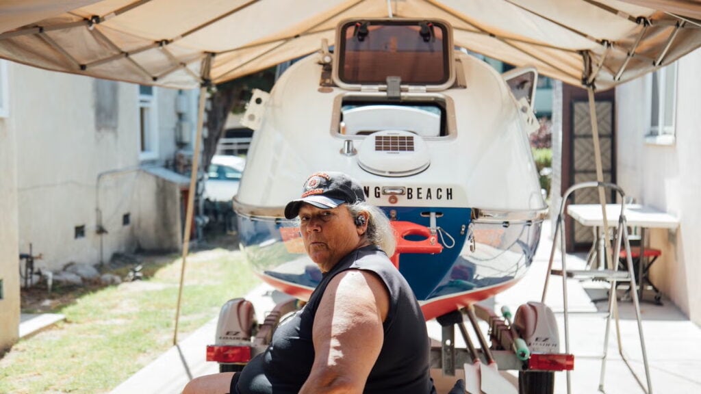 a white woman wearing a baseball cap over curly gray hair, in front of a rowing boat on a trailer in a grassy yard. 