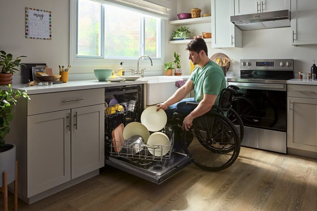 Man using a manual wheelchair in a kitchen loads a plate into a full disherwasher rack, that is turned slightly sideways. 