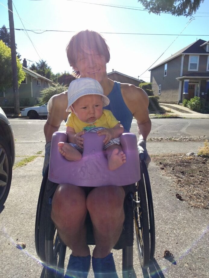 male manual wheelchair user outside with a rubber bumbo seat and a baby on his lap.