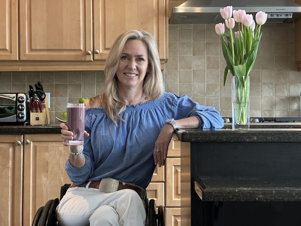 A blond woman holding a smoothie, using a manual wheelchair and sitting in her kitchen.