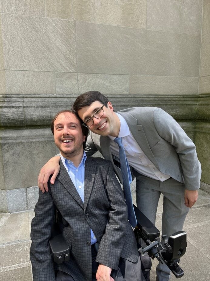 two men wearing suits one is sitting inn a power wheelchair, the other is bending over with a hand around the others shoulder. 