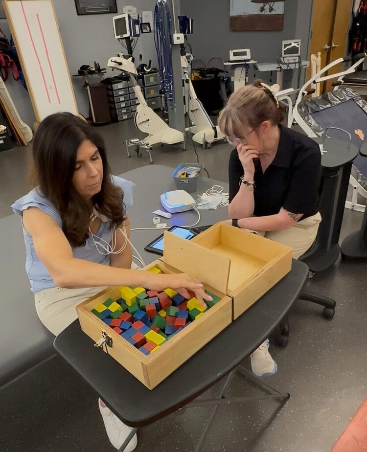 two women sitting in a clinical setting. One is picking colored blocked out of a box.
