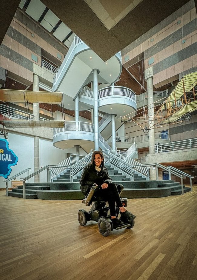 woman using a power wheelchair sitting in a museum atrium with early wood and fabric airplanes hanging from the ceiling.