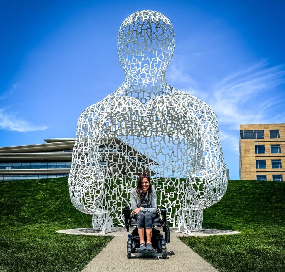 woman using a power wheelchair sits on a paved path in front of a steel sculpture