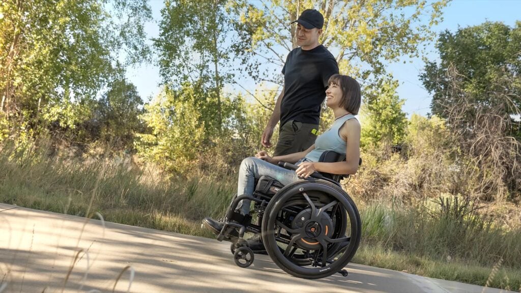 A woman in a manual wheelchair with power wheels rolling outside on a paved path with a man walking next to her. 