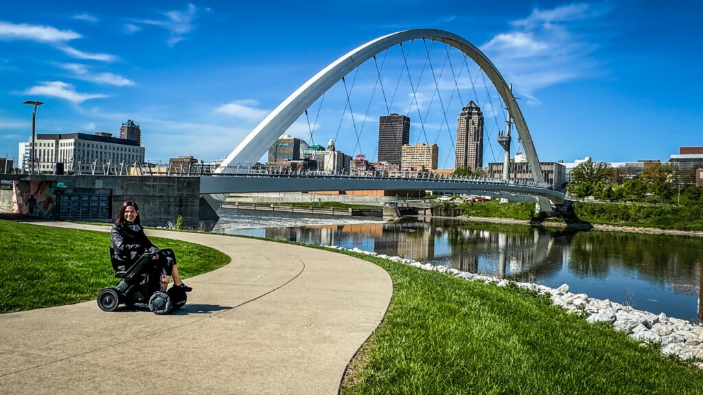 a woman using. apower wheelchair sits on the grass in front of a large steel sculpture. 
