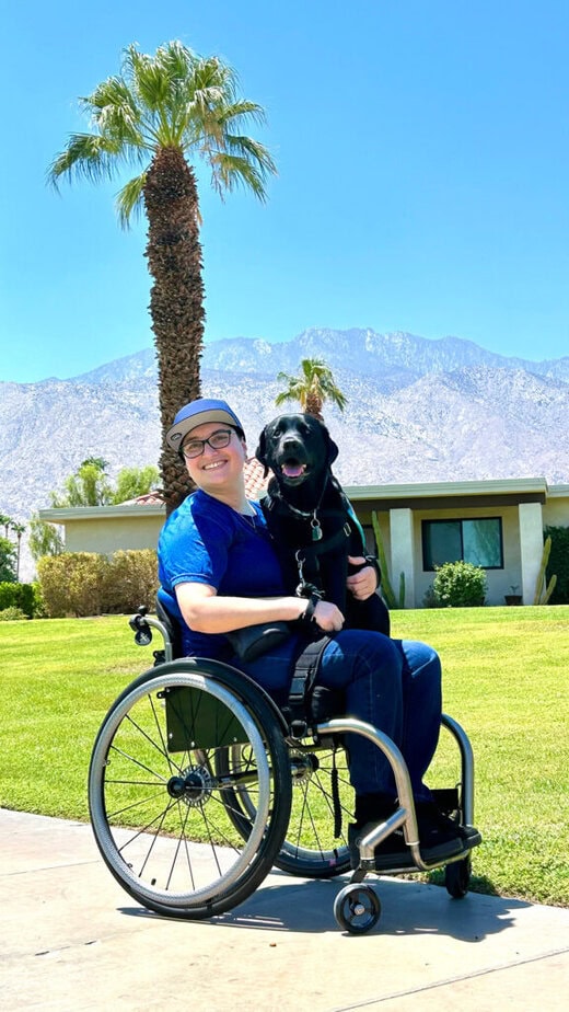 A woman in a wheelchair smiling outdoors with her service dog in front of a sunny house and mountain backdrop.